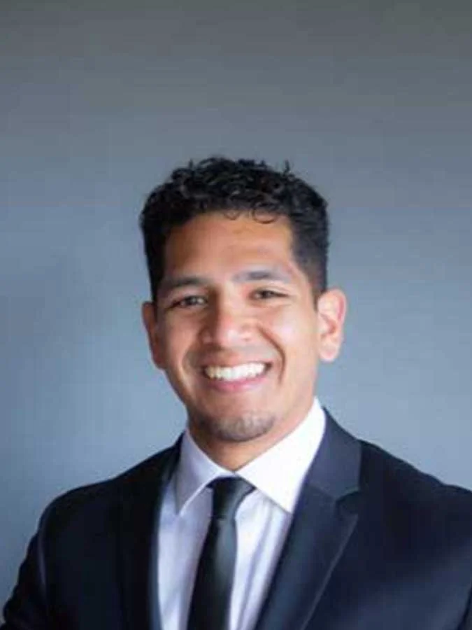 A man with short curly hair and a trimmed beard smiles while wearing a black suit, white shirt, and black tie against a plain gray background.