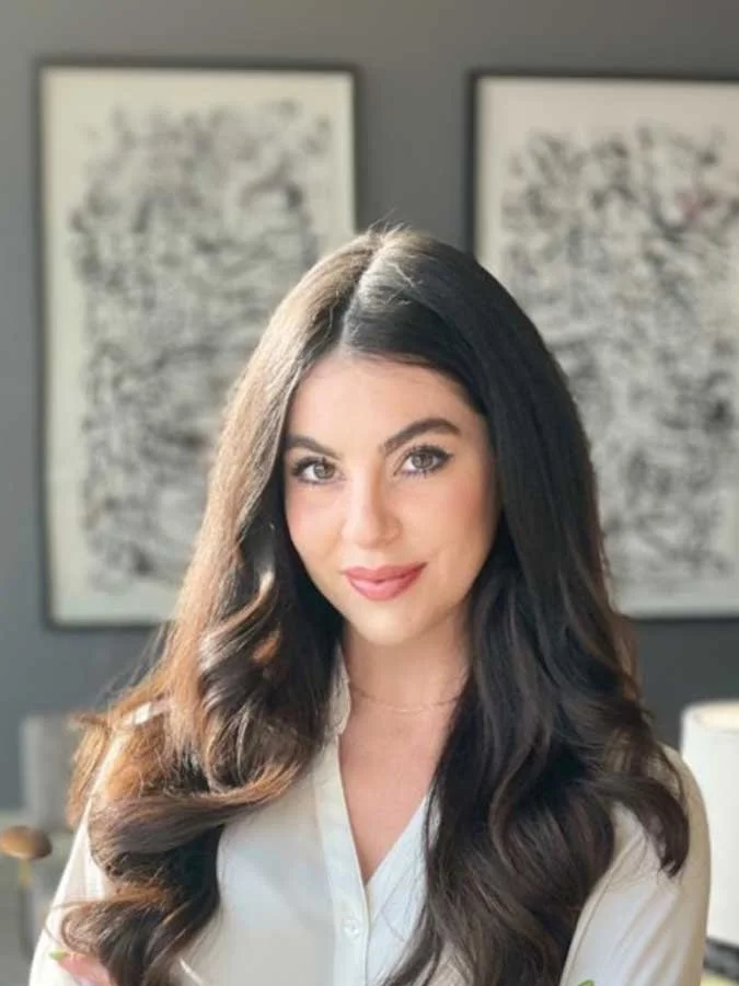 A woman with long dark hair and a white blouse smiles at the camera, standing indoors with two abstract black and white artworks on the wall behind her.